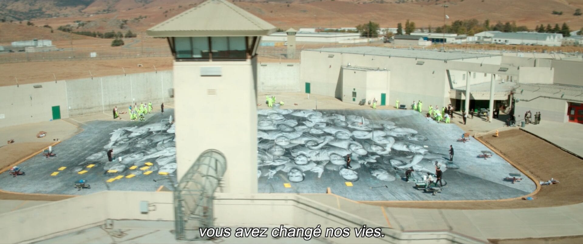 a still from a documentary in a prison in California showing inmates working on installing a large mural of portraits of themselves in the prison yard